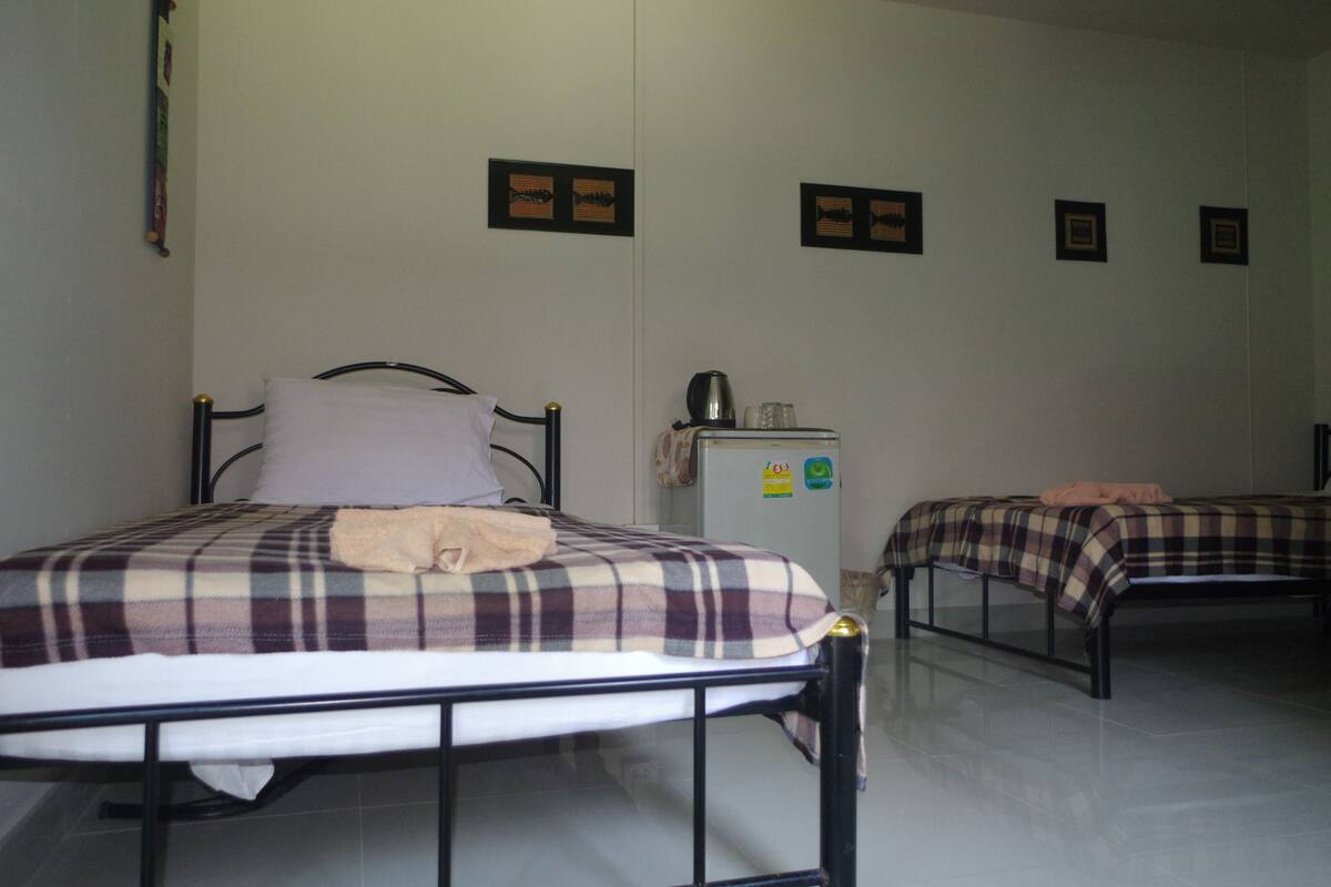 A room with two beds, each with a plaid bedspread and a pillow. There is a small refrigerator with magnets and a kettle on top.