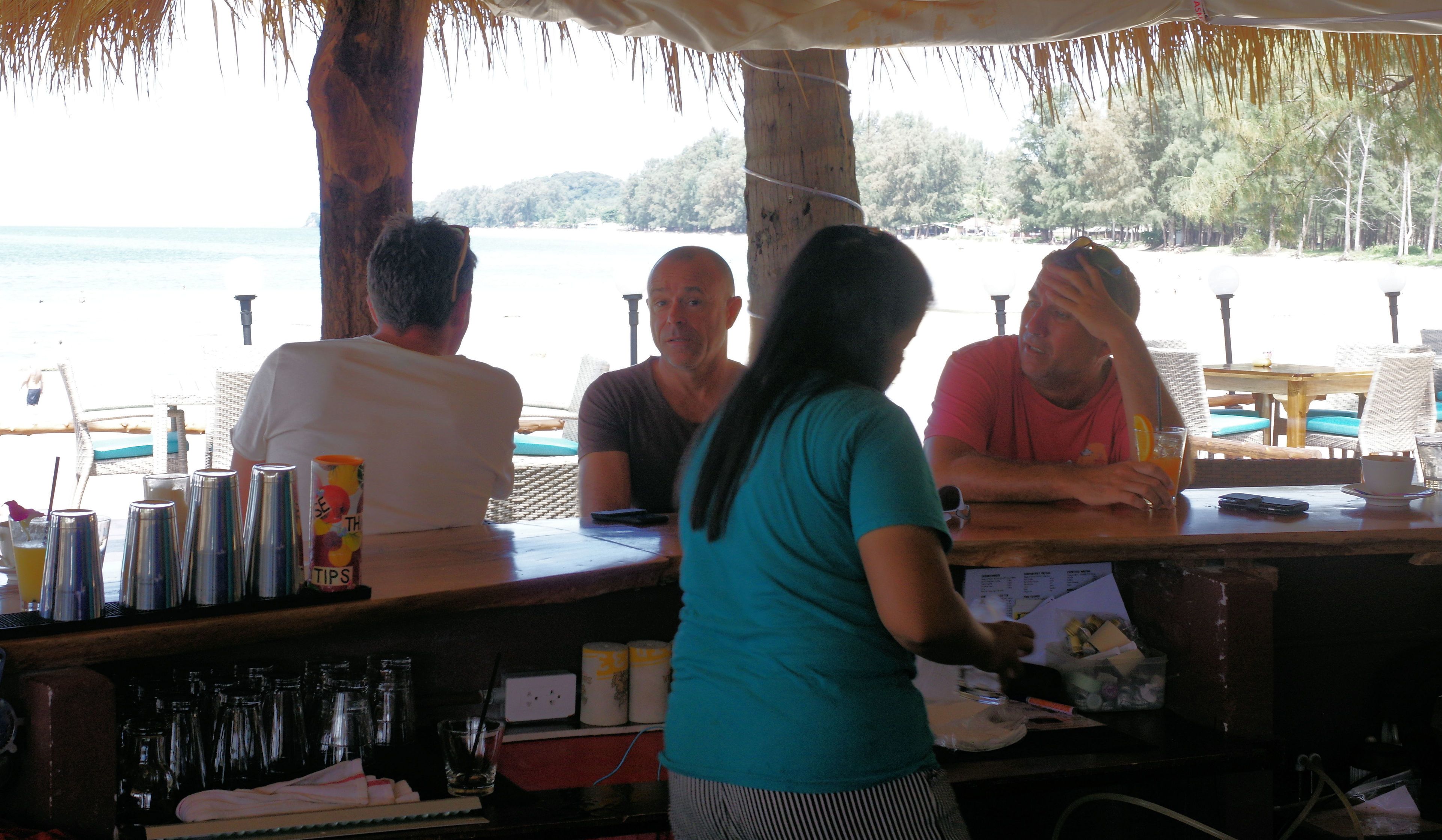 A bar area with a view of the beach, wooden furniture, and a person serving drinks.