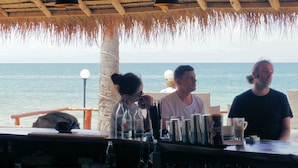 Un bar en la playa con techo de paja, barandilla de madera y vista al mar.