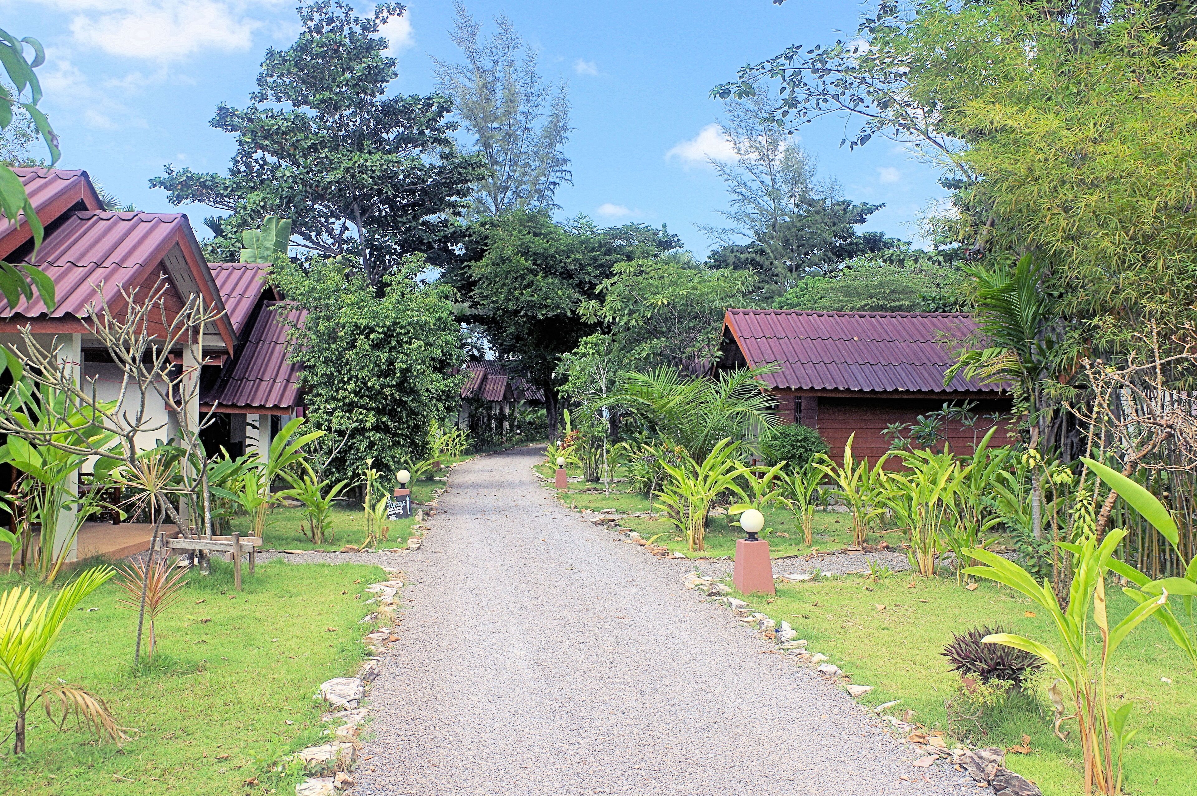A pathway leading to a row of rustic-style buildings with red roofs, surrounded by lush greenery and trees.