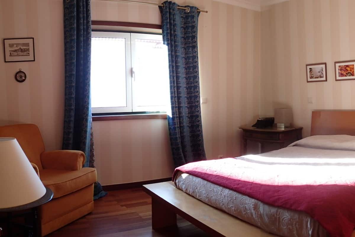 Une chambre à coucher avec un lit, une chaise, une table de chevet, une lampe et une fenêtre avec des rideaux.