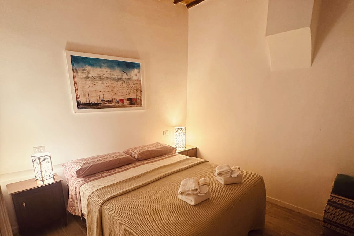 Un dormitorio con techo de madera, una cama con manta a rayas, una mesita de noche con lámpara y un cuadro en la pared.