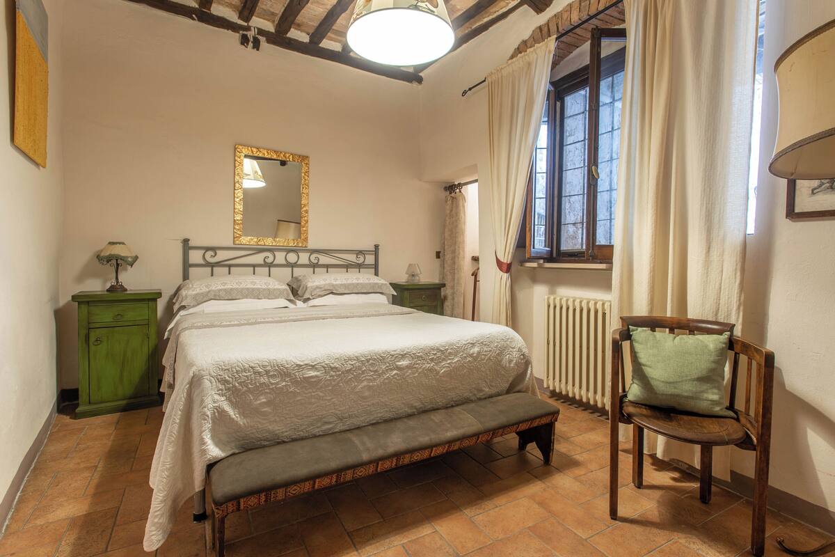 Un dormitorio con cama, una silla, mesita de noche, cómoda, ventana con cortinas, radiador y un espejo en la pared.