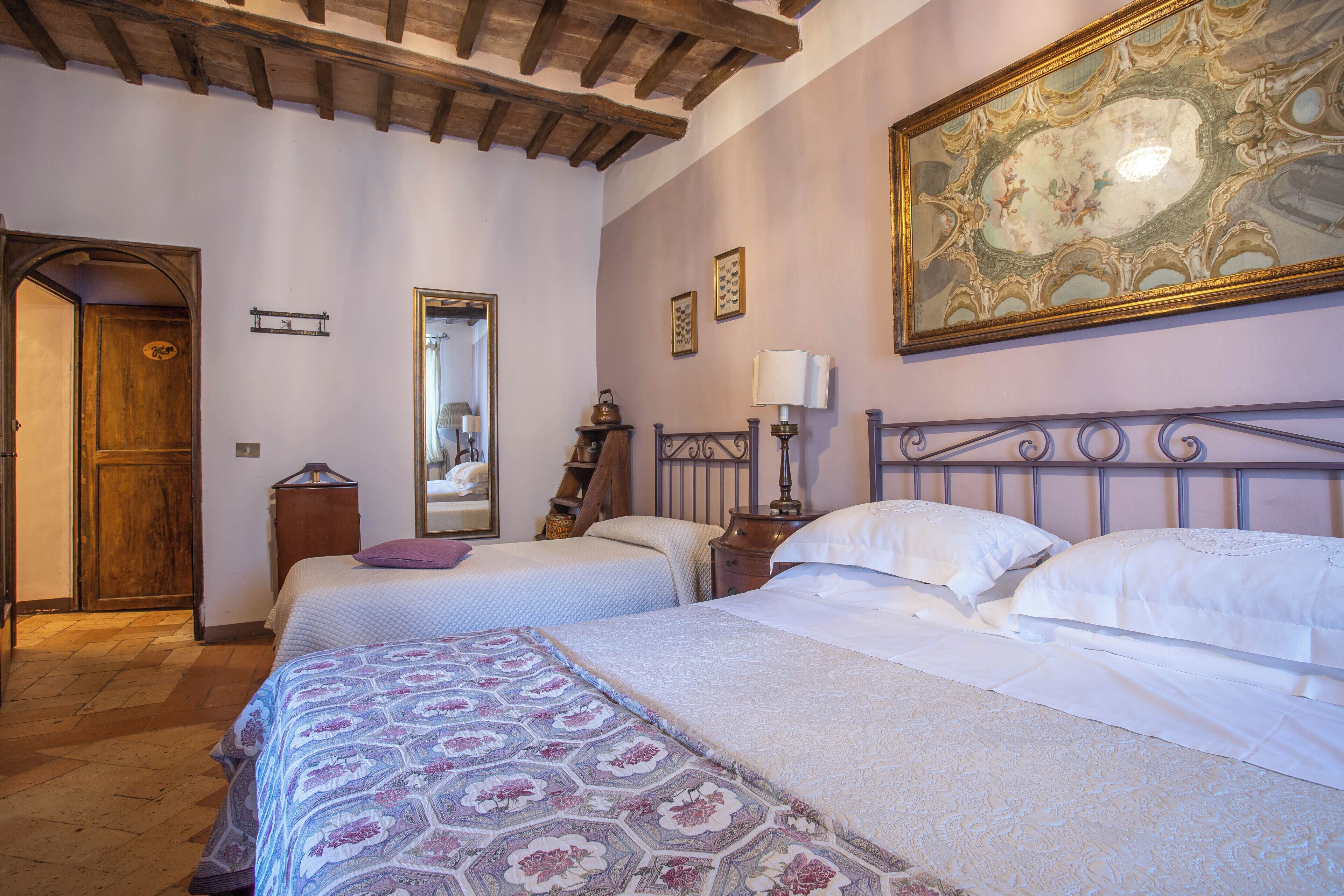 Une chambre avec deux lits, des poutres en bois au plafond, un grand miroir décoratif et une fresque murale.
