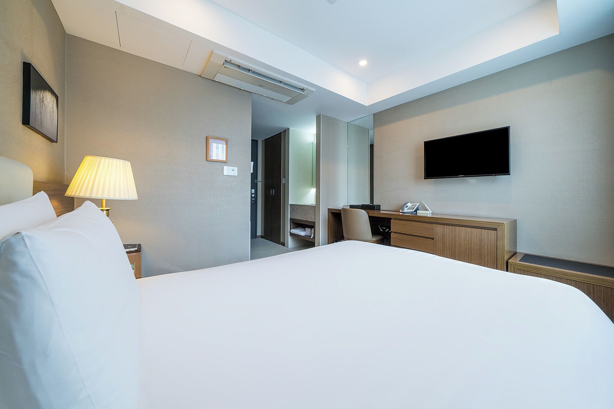 A hotel room with a large bed, a desk with a chair, a flat-screen TV, and a lamp.