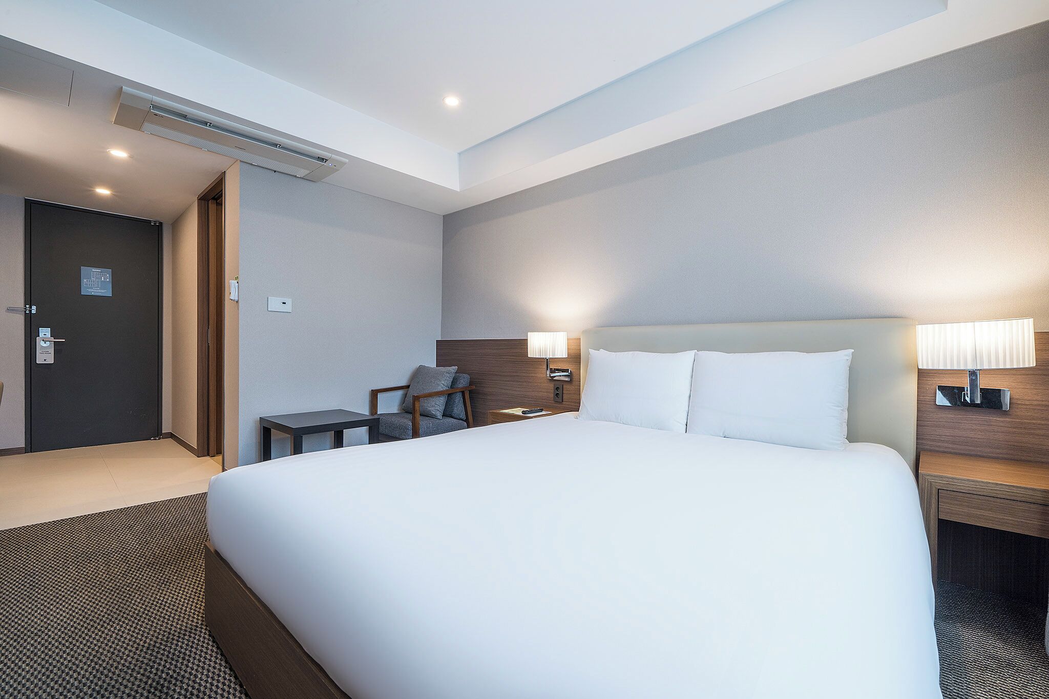 A hotel room with a large bed, a desk, a chair, a nightstand, and a wall-mounted lamp.