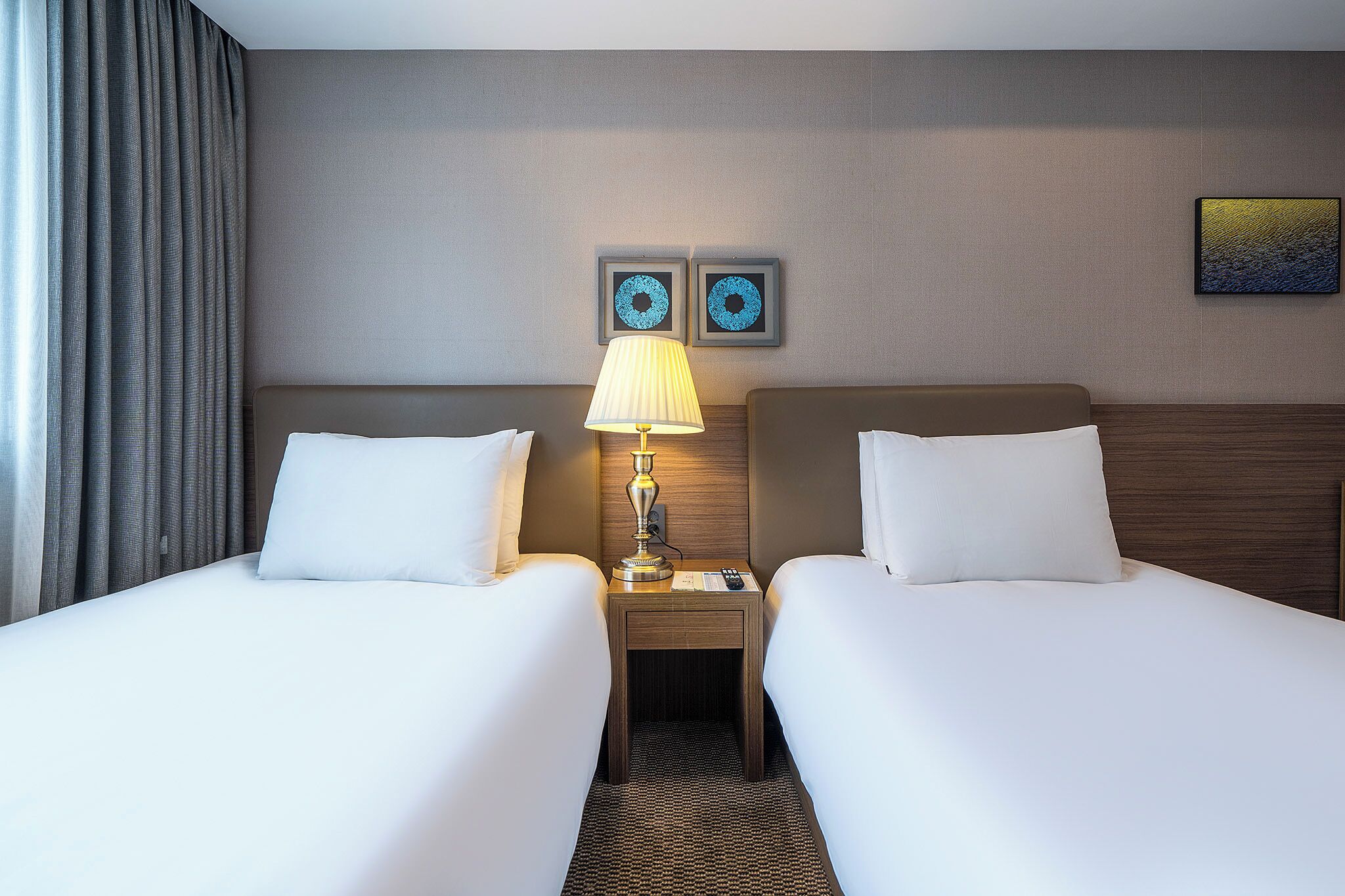 A hotel room with two beds, a nightstand with a lamp, and two wall art pieces.