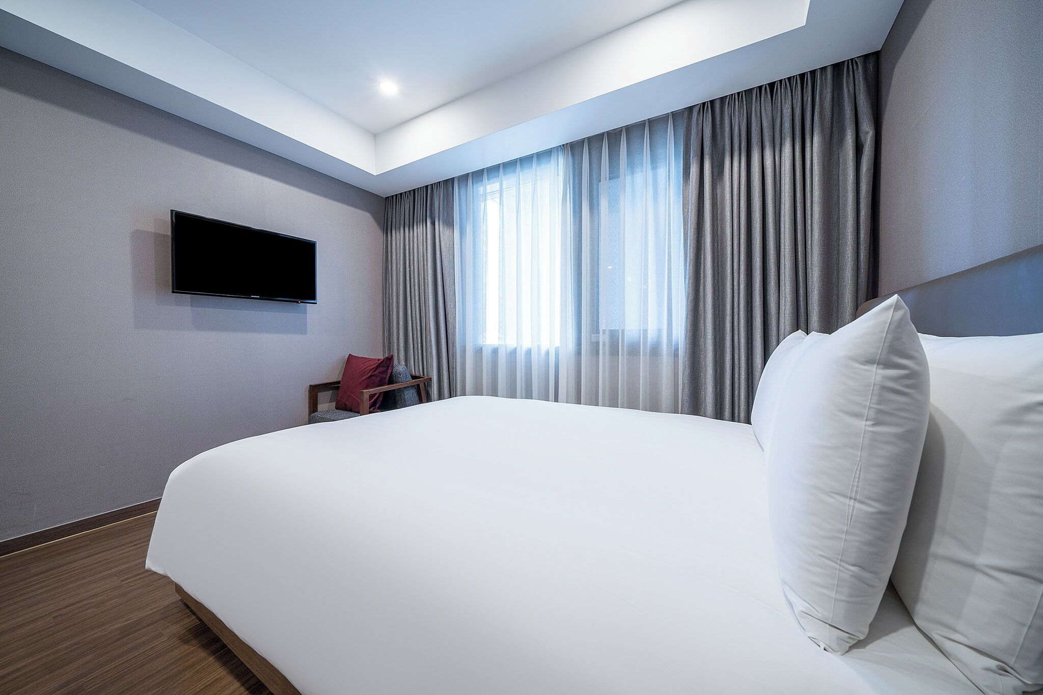A hotel room with a large bed, a flat-screen TV mounted on the wall, a window with sheer curtains, and a wooden floor.