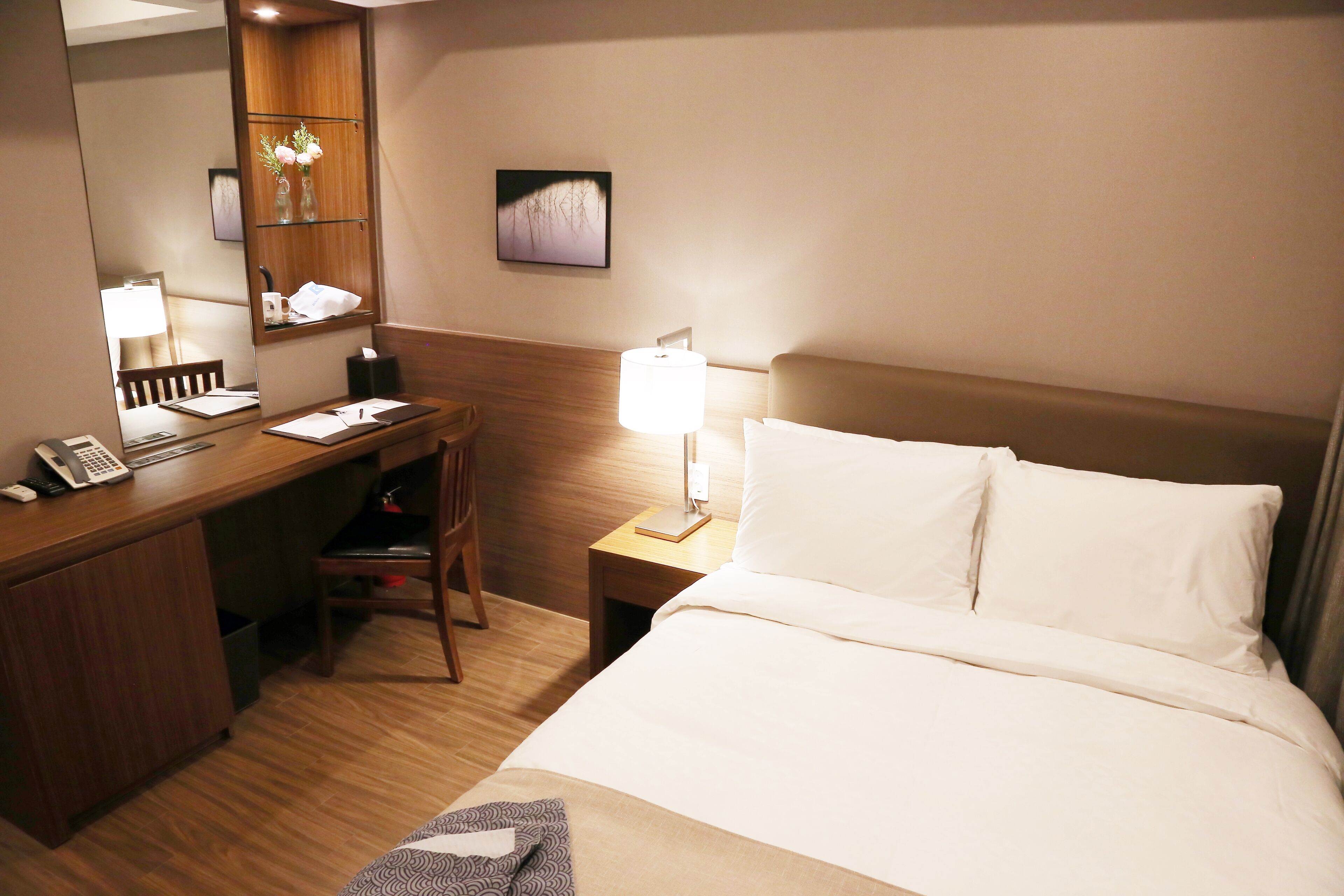 Une chambre d’hôtel avec un bureau en bois, une chaise, un lit avec du linge de lit blanc, une lampe de chevet, une petite table avec un vase de fleurs et un tableau encadré au mur.