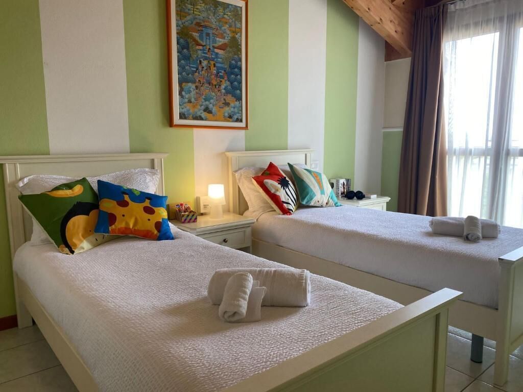 A hotel room with two beds, each with a white bedspread and colorful pillows. There is a painting on the wall above the beds and a window with curtains.