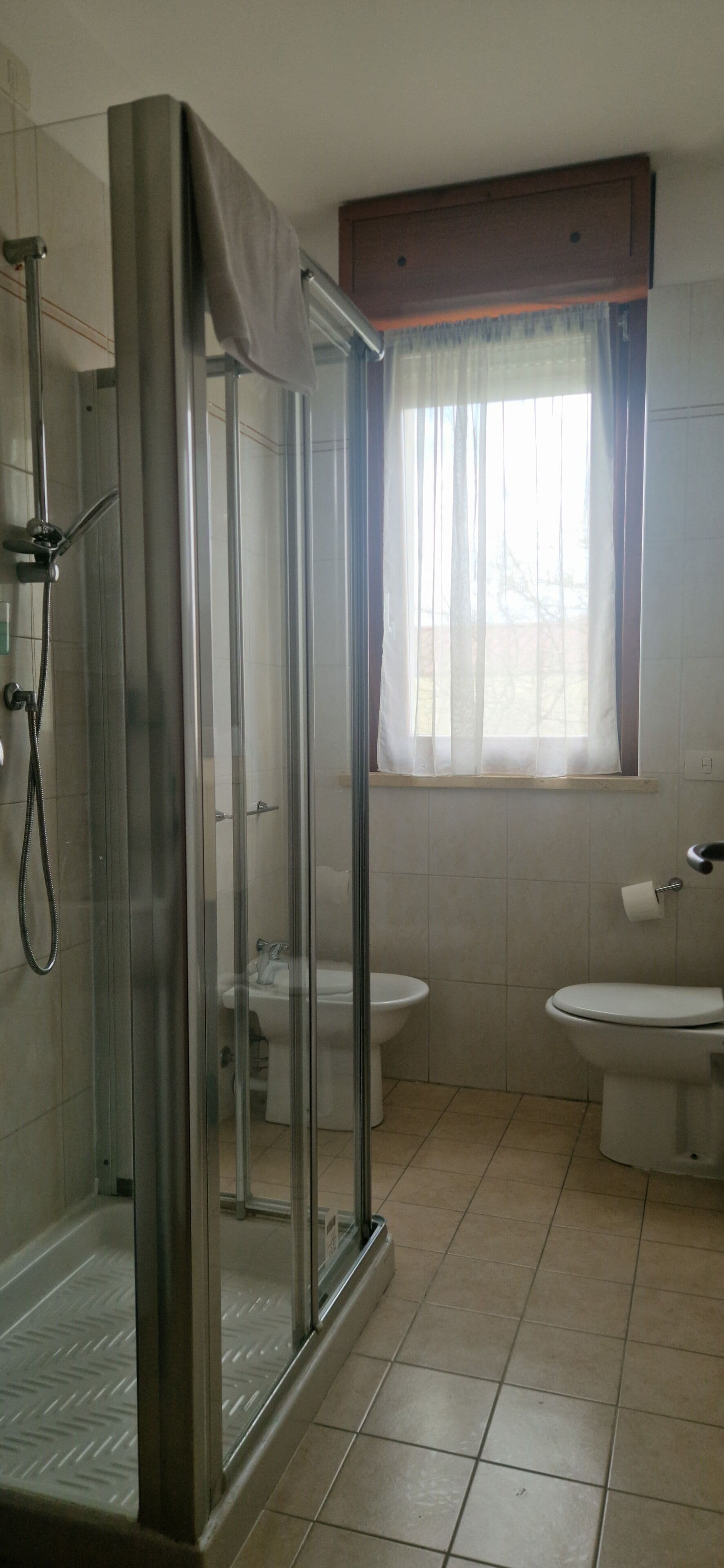 A bathroom with a glass-enclosed shower, two toilets, a sink, and a window with curtains.