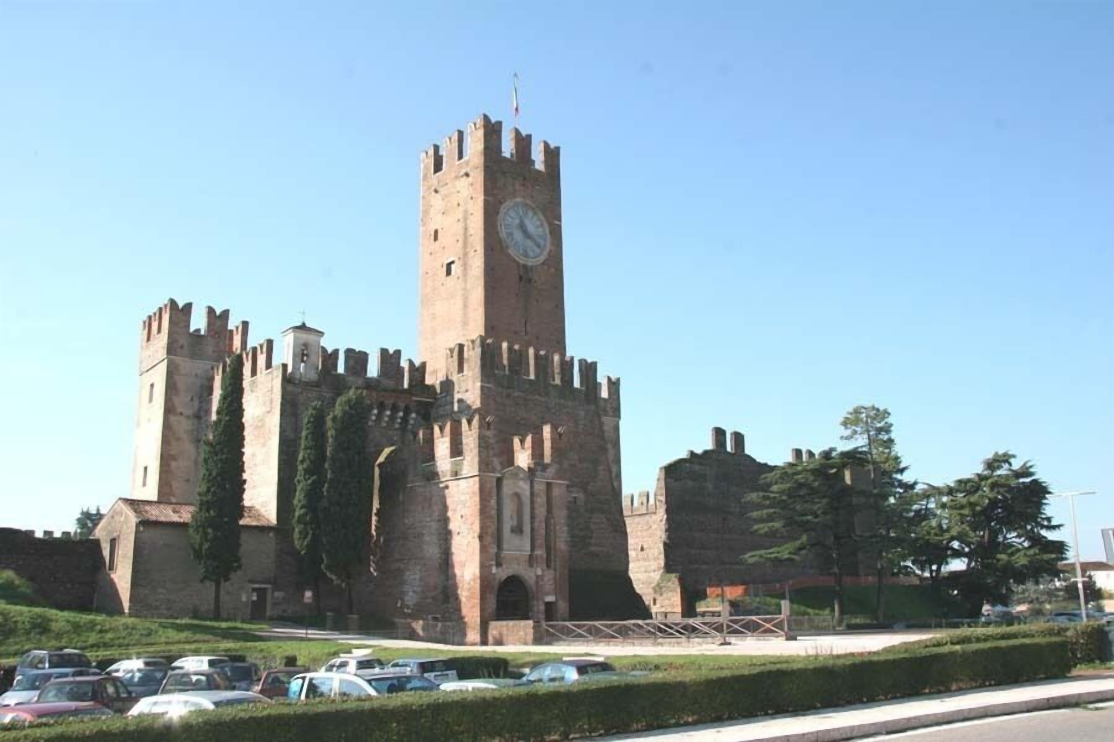 Un castillo histórico con una imponente torre del reloj, rodeado de aparcamiento y vegetación.