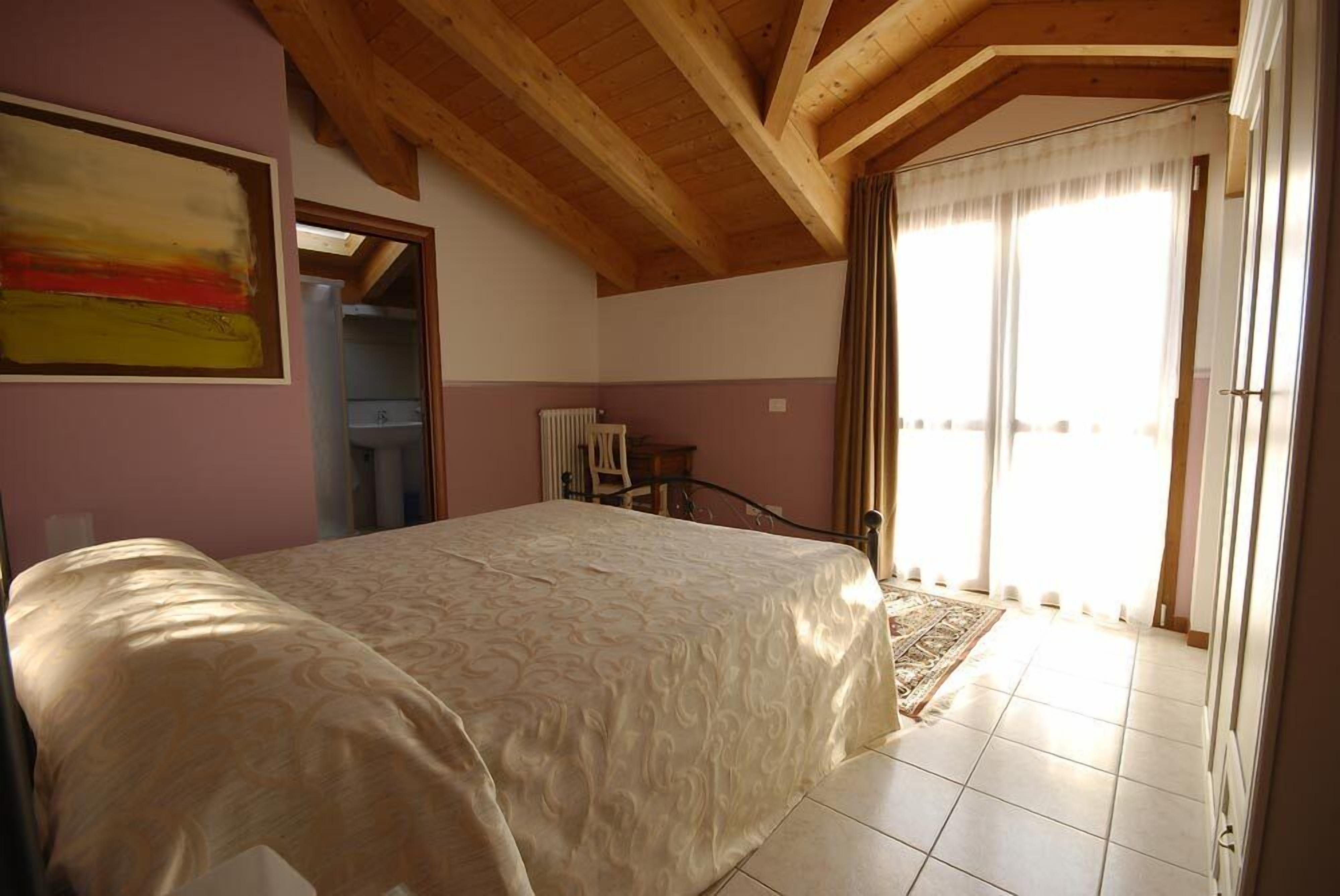 A bedroom with a large bed, wooden ceiling beams, a radiator, and a view of the outside through a door.