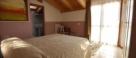 A bedroom with a large bed, wooden ceiling beams, a radiator, and a view of the outside through a door.