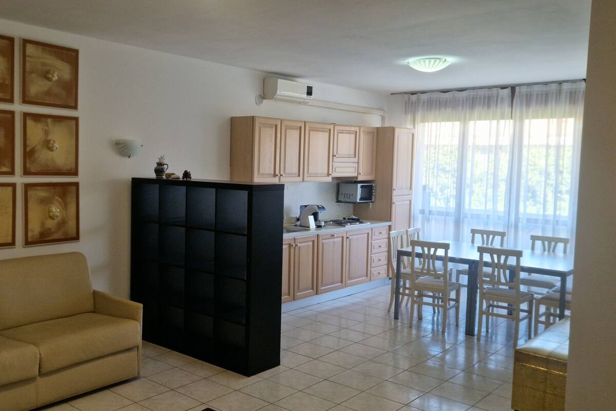A living room with a sofa, a black cabinet, a dining area with chairs, a kitchen with cabinets and appliances, and a large window with curtains.