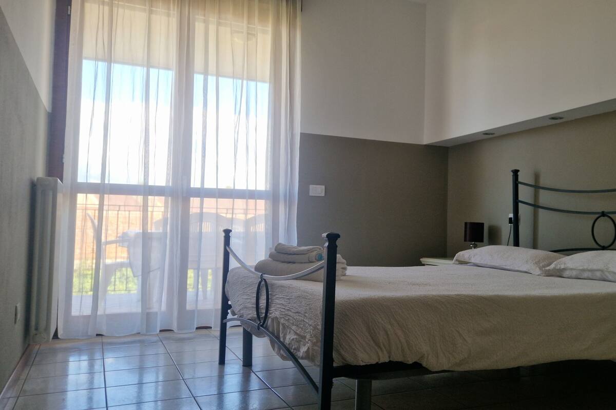 A bedroom with a bed, a nightstand, a window with curtains, and a view outside.