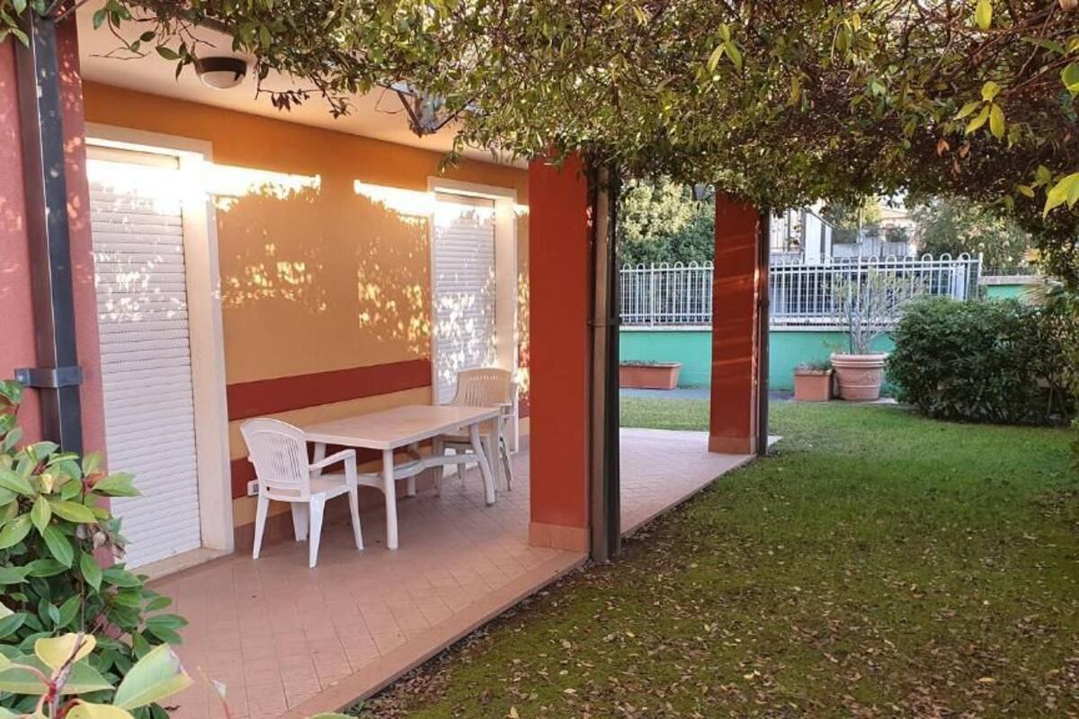 A covered patio area with a table and chairs, surrounded by greenery and a fence.