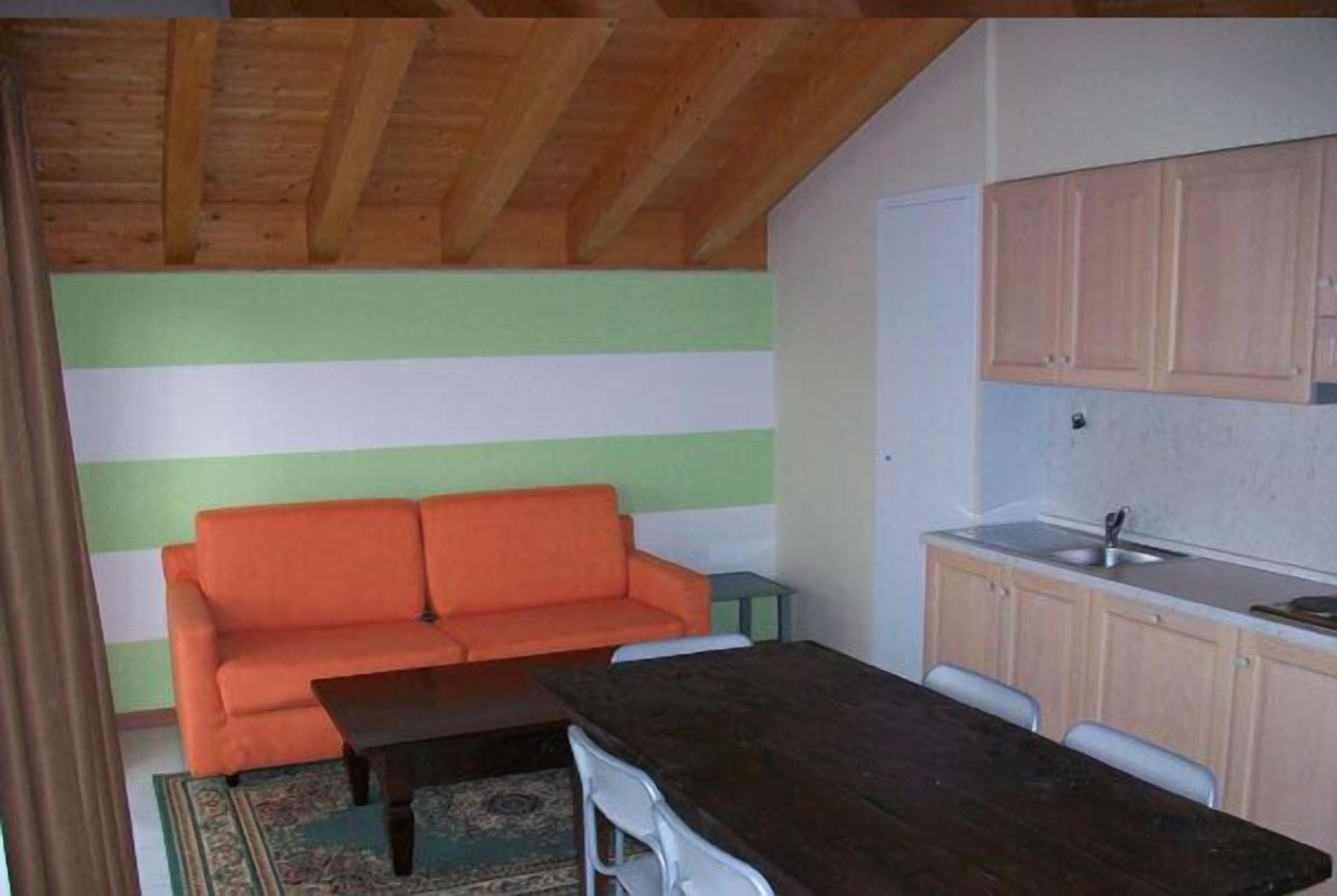A compact room with a wooden ceiling, a green and white striped wall, a small orange sofa, a dark wooden table, and a kitchen area with light wooden cabinets and a sink.
