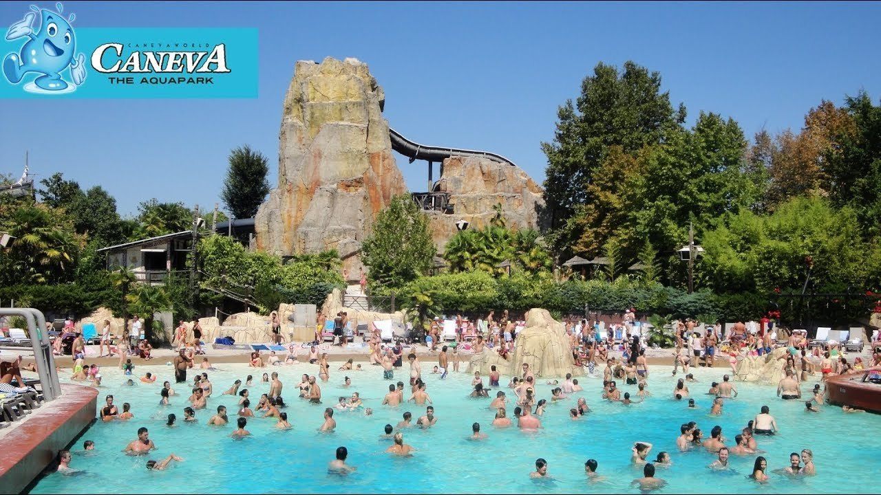 Una amplia piscina al aire libre con gente disfrutando del agua, rodeada de árboles y una estructura rocosa al fondo.