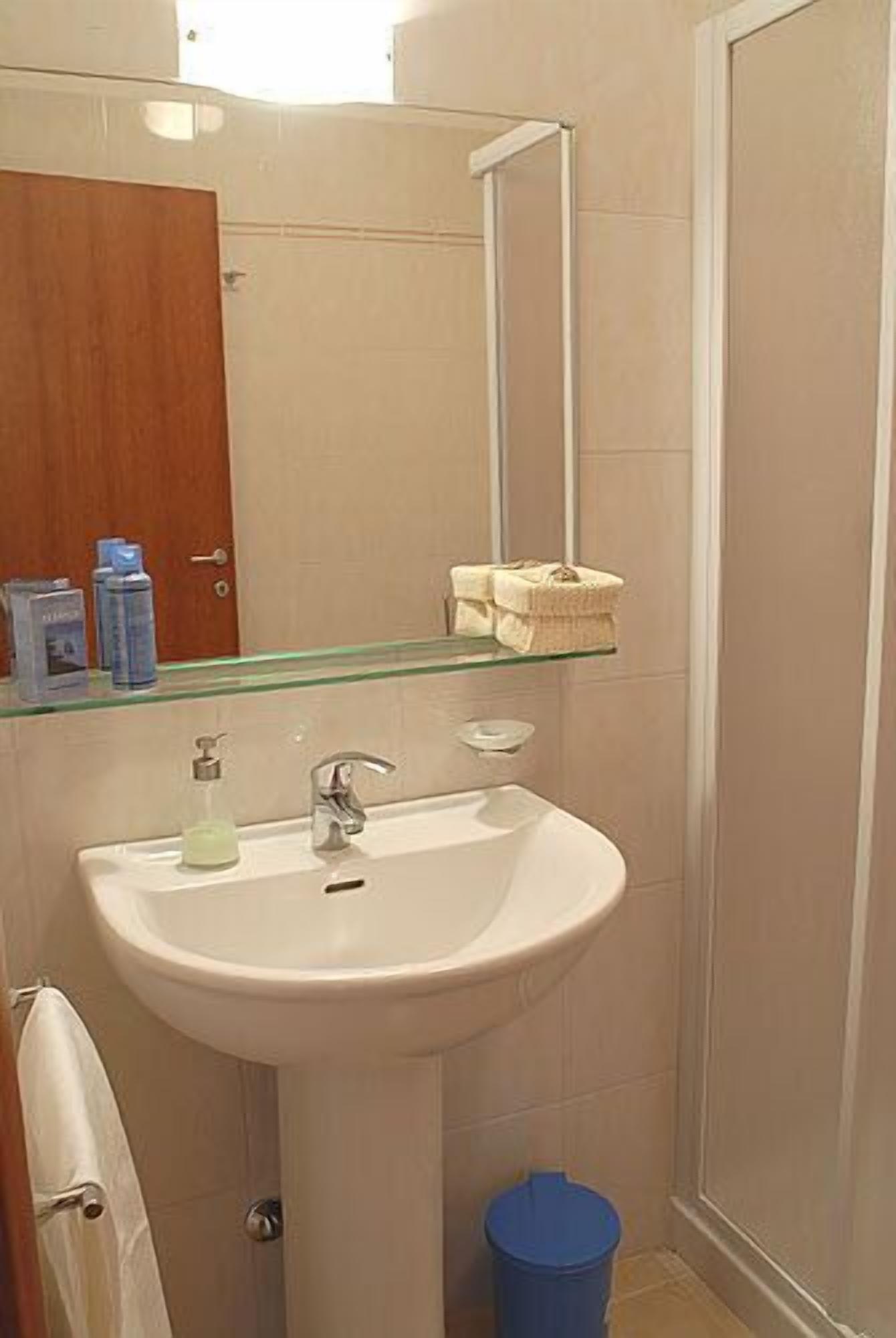 A bathroom with a white sink, a glass shelf above the sink, a mirror, and a blue trash bin.