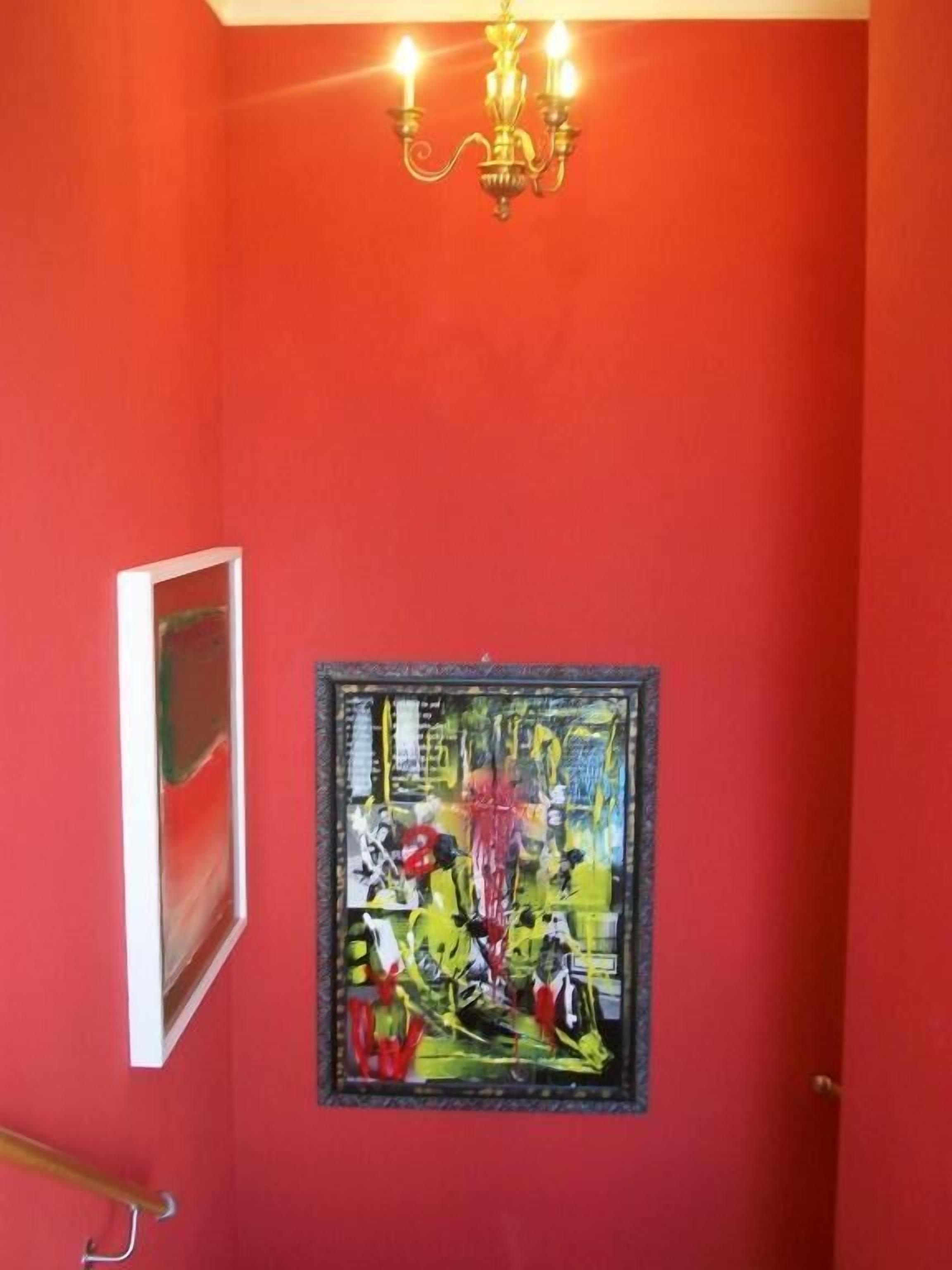Una habitación con una pared roja, un cuadro abstracto enmarcado, una puerta blanca y un candelabro.