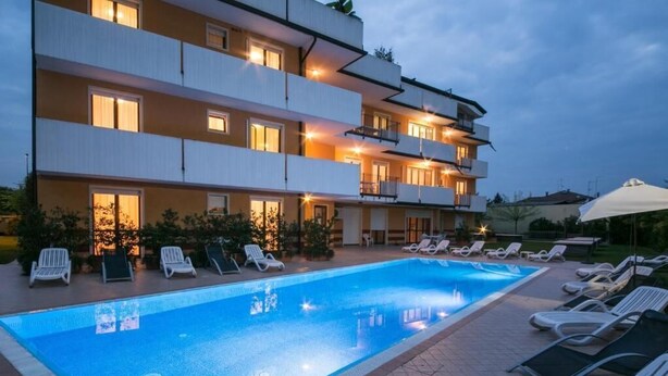 A swimming pool with lounge chairs and a multi-story building with balconies. - Residence Garda Palace  (Peschiera del Garda)