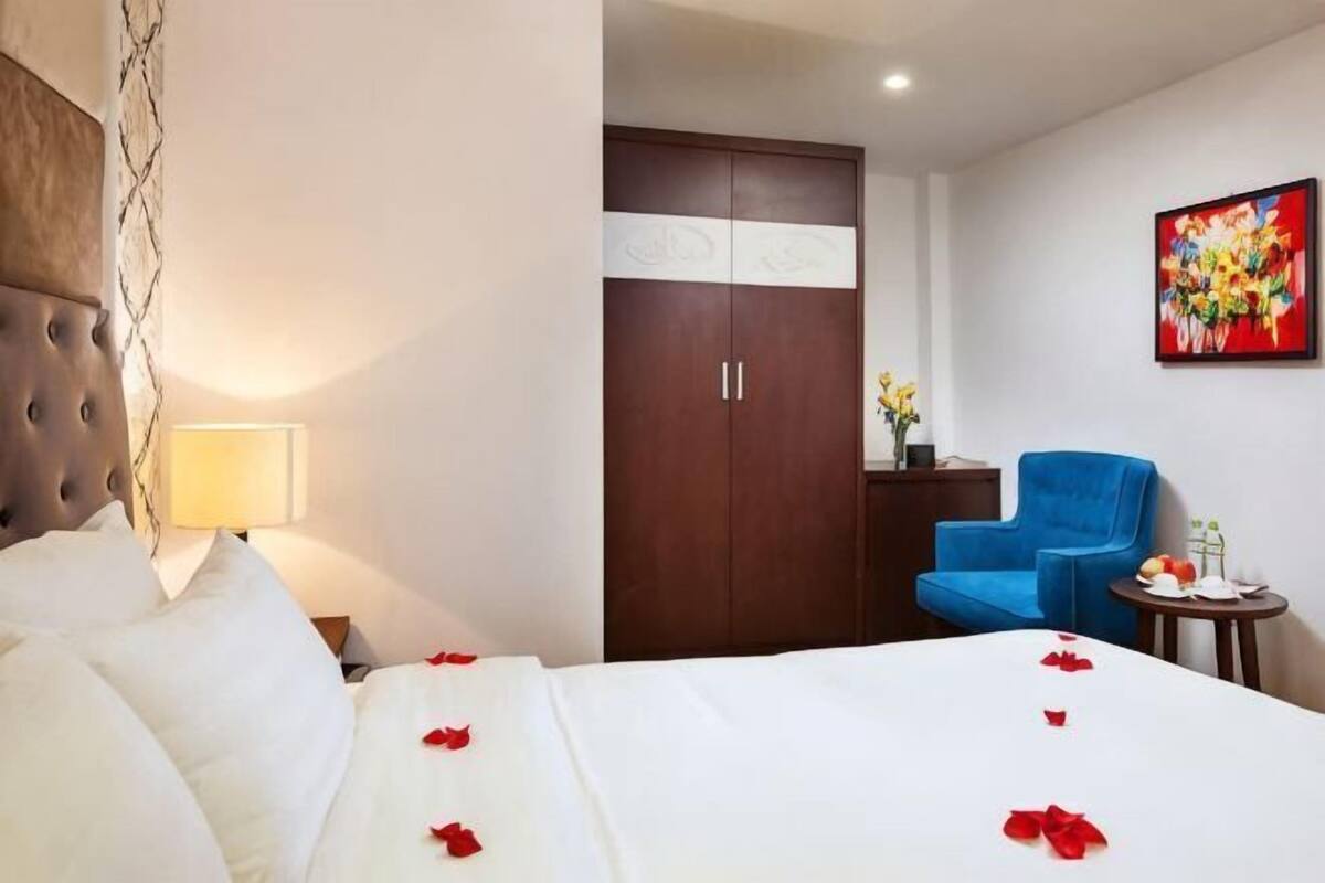 A hotel room with a bed, a wardrobe, a blue armchair, and a table with a vase of flowers.