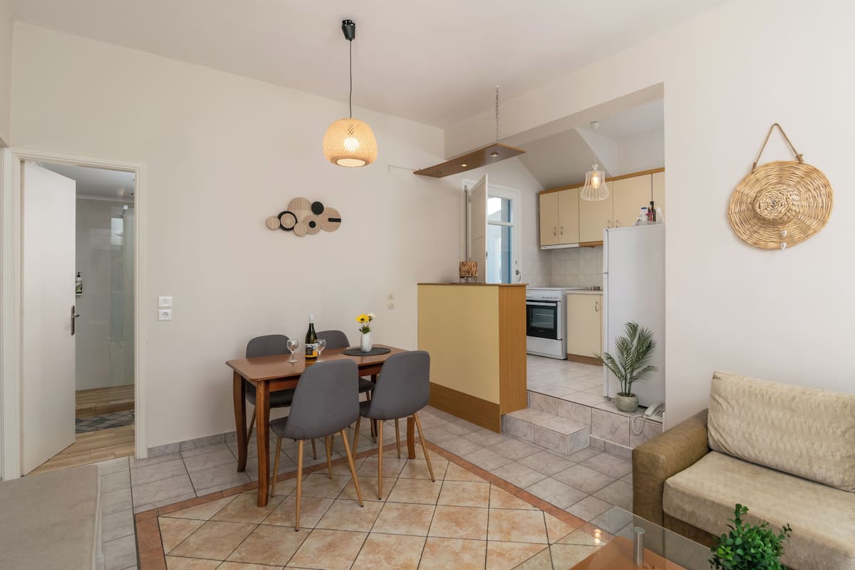 A modern kitchen with a central island, a dining area with a table and chairs, and a living area with a sofa and a plant.