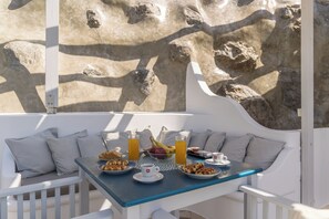 Un área de estar exterior blanca con una mesa dispuesta para el desayuno, incluyendo platos de comida, un frutero y bebidas.