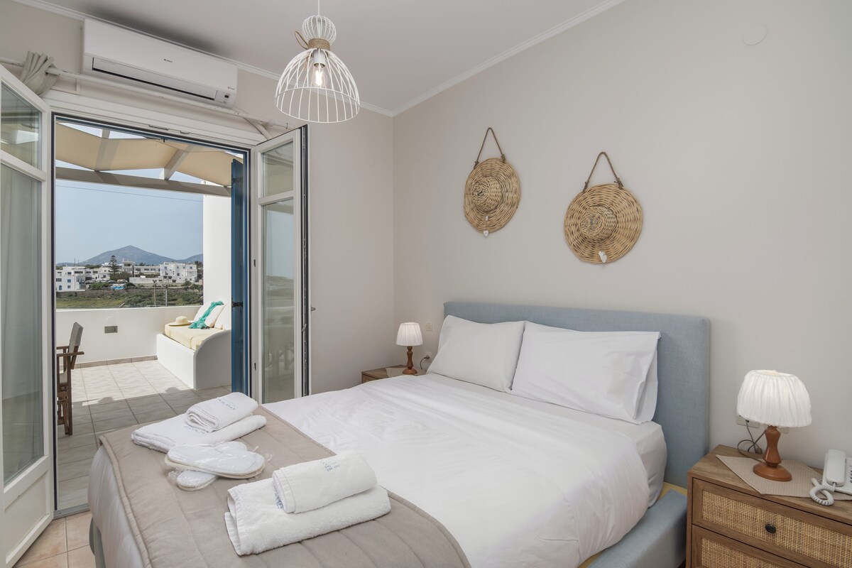 A hotel room with a bed, bedside table, lamp, and a view of the outside.
