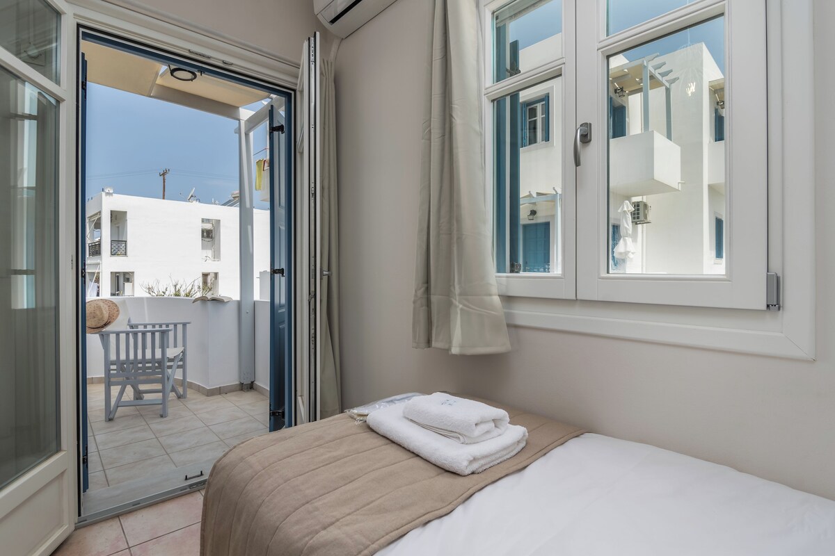 Een hotelkamer met een bed, een raam met gordijnen, een balkon met een stoel en uitzicht op gebouwen.