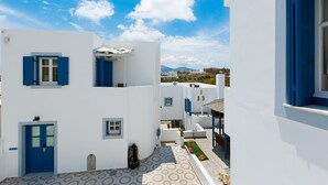 A white building with blue shutters and a door, a small garden area, and a view of distant hills. -  Vivere Houses (Milos)