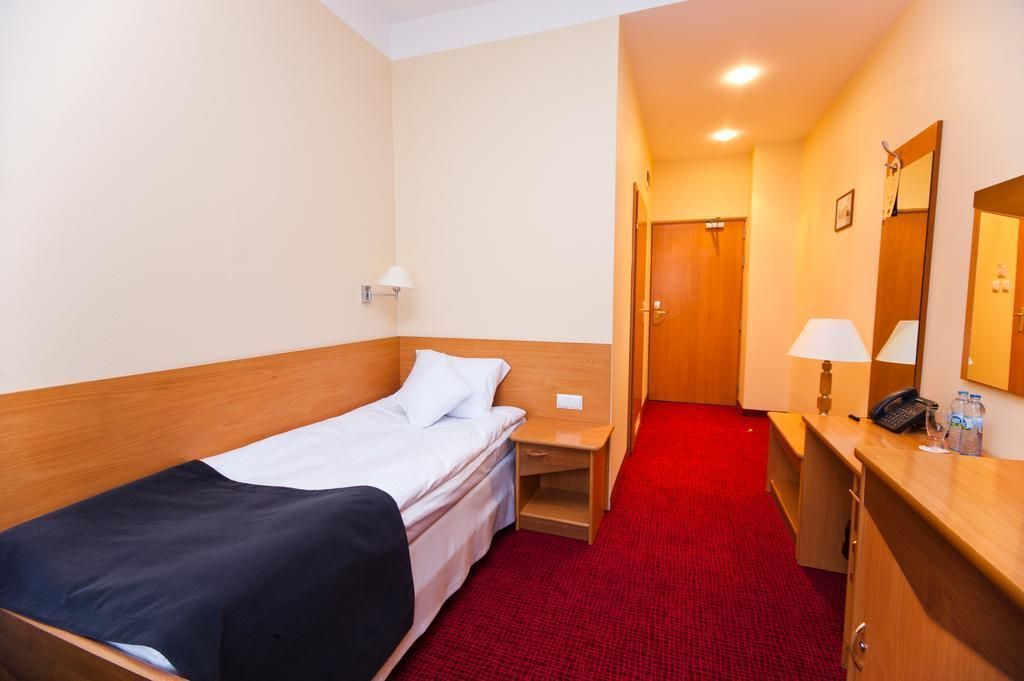 A hotel room with a single bed, a bedside table with a lamp, a desk with a mirror, and a red carpeted floor.