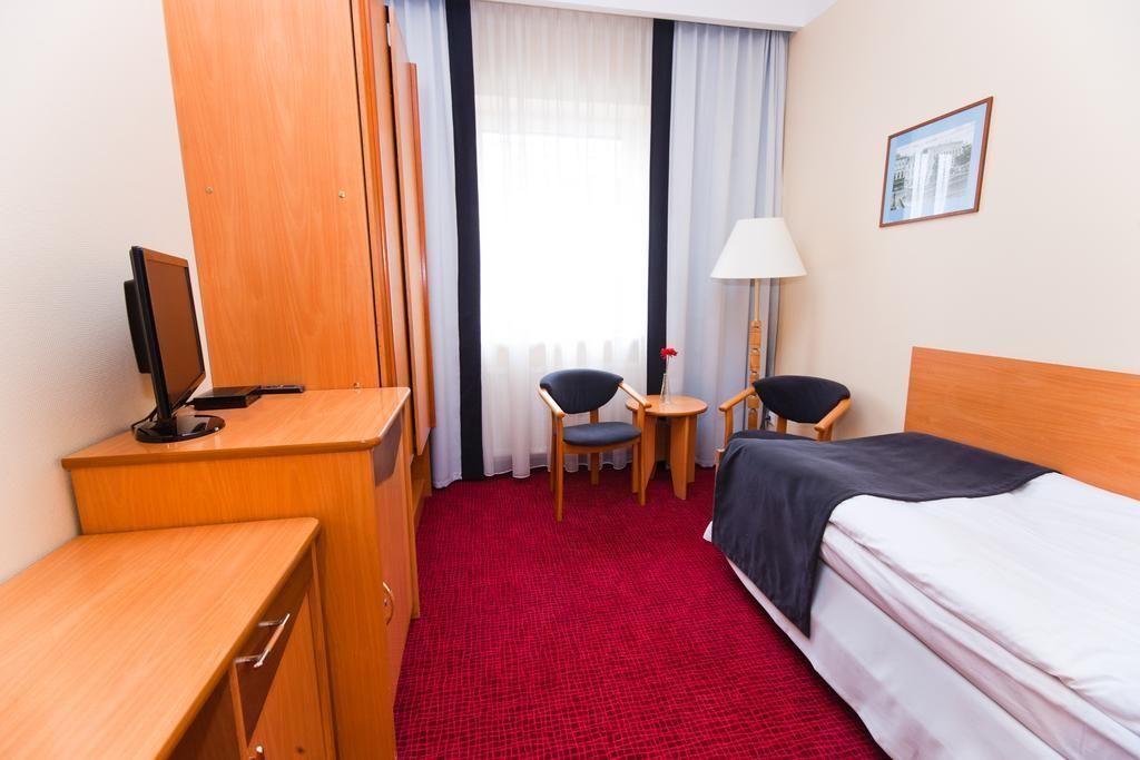 A hotel room with a bed, a desk with a computer, two chairs, a lamp, and a window with curtains.
