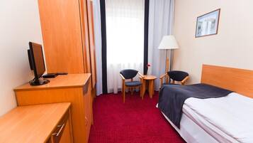 A hotel room with a bed, a desk with a computer, two chairs, a lamp, and a window with curtains.
