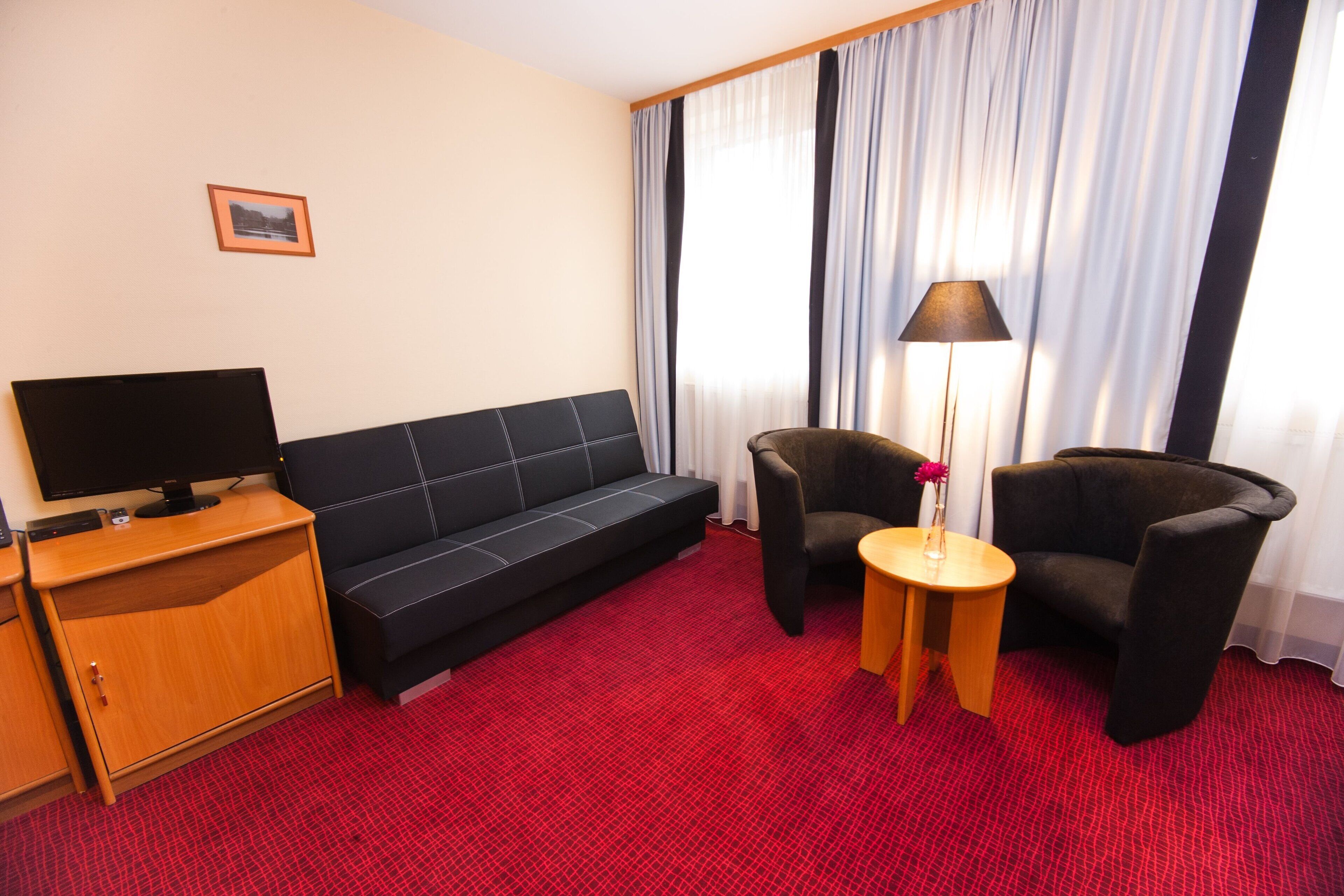 Habitación de hotel con sofá negro, dos sillones, un televisor sobre un mueble de madera y una lámpara en una mesita.