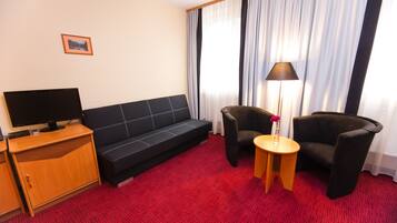 A hotel room with a black sofa, two armchairs, a TV on a wooden cabinet, and a lamp on a small table.