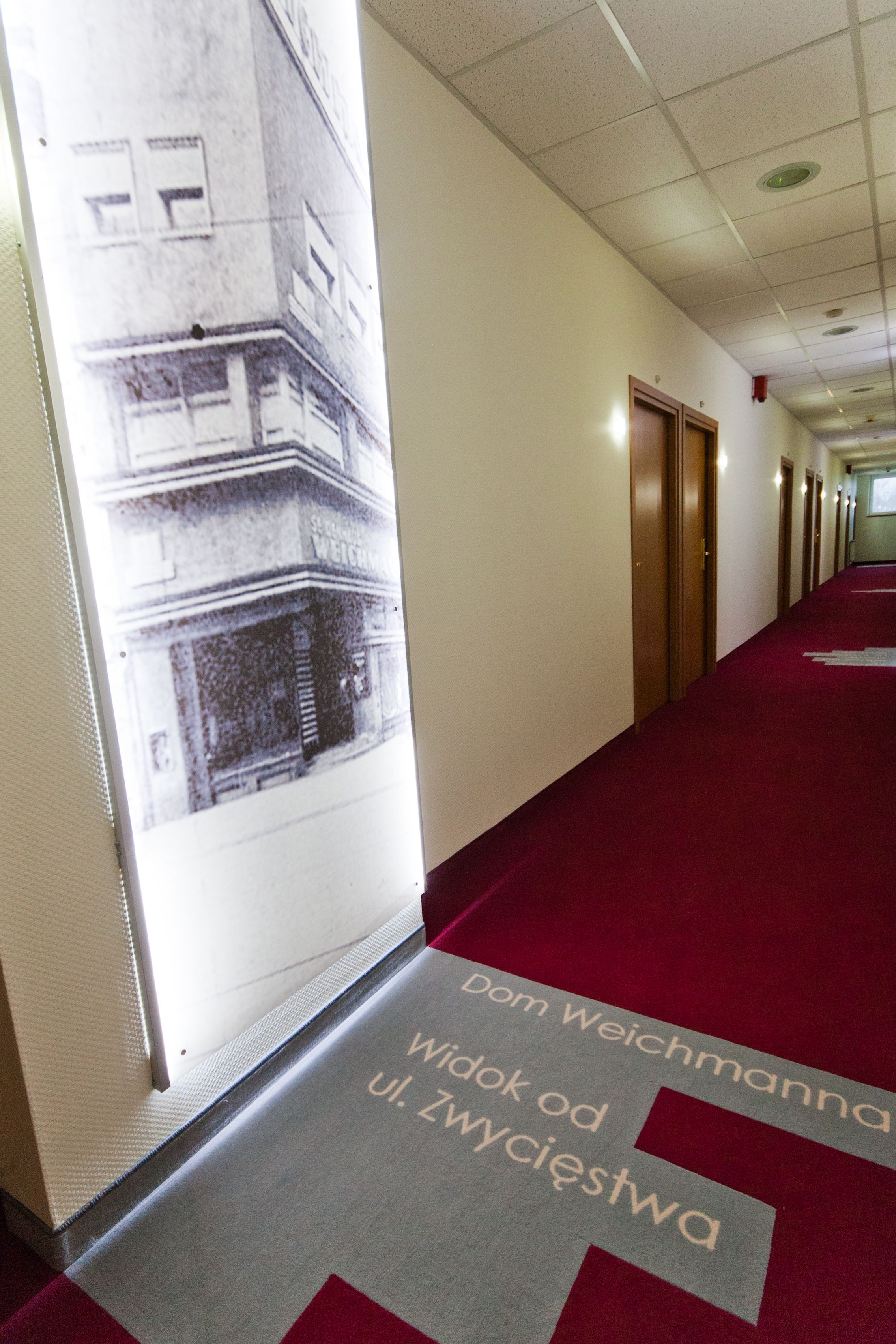 Un couloir d’hôtel avec une moquette rouge et une grande affiche accrochée au mur.
