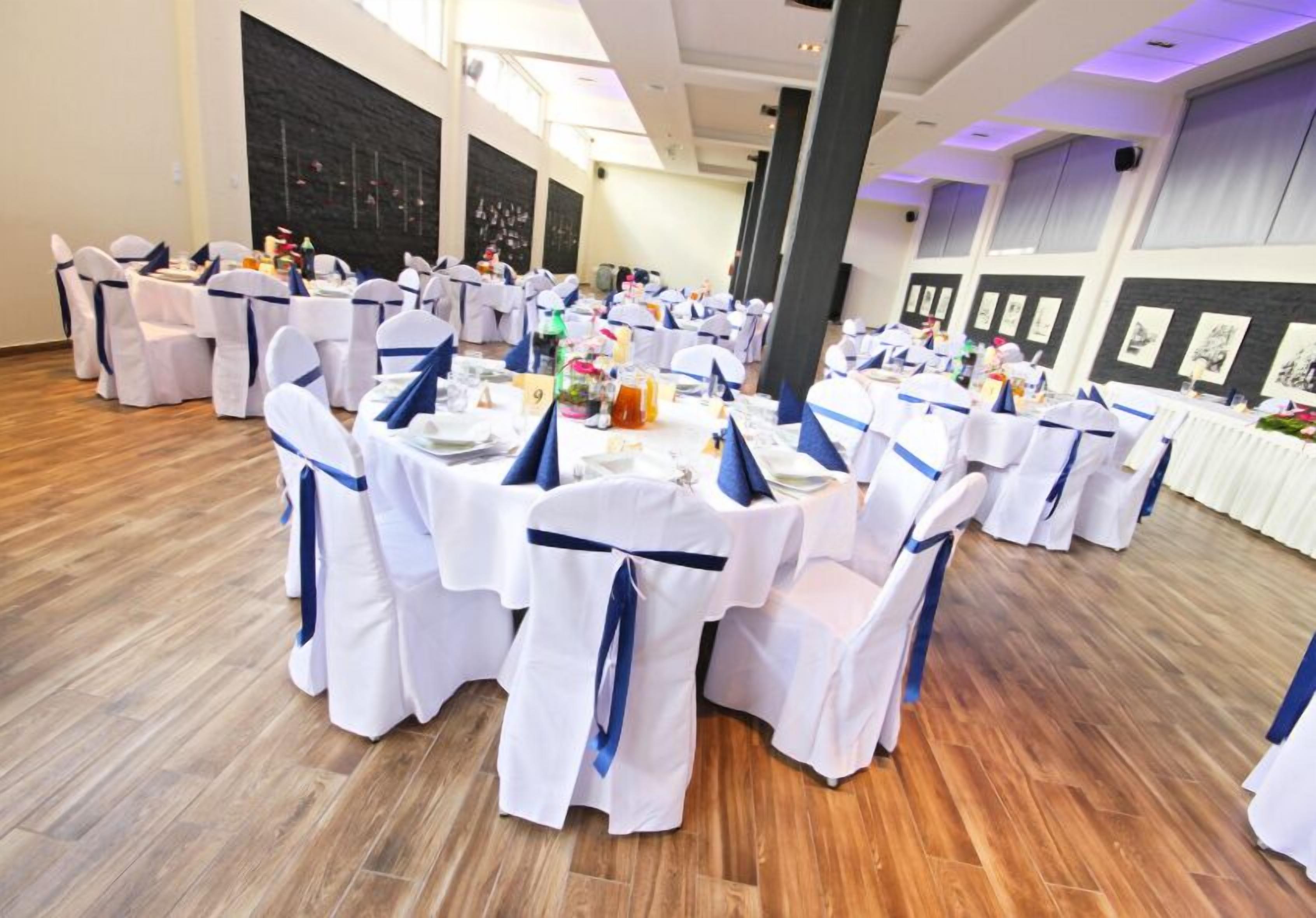 A banquet hall with round tables covered in white tablecloths, each with blue sashes. The room has wooden flooring and large windows allowing natural light.