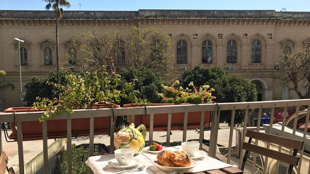 A balcony with a table set for a meal, potted plants, and a building with arched windows in the background. - Nuvole Barocche (Lecce)