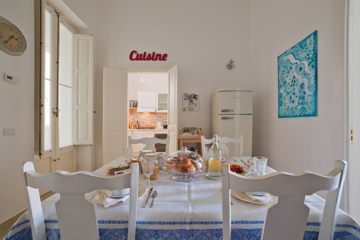 A dining area with a table set for two, a kitchen in the background, and a clock on the wall.