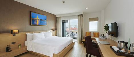 A hotel room with a large bed, a desk with a laptop, a television, and a balcony with a view of buildings.