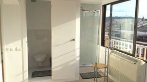 A small, bright room with a toilet, a shower, and a radiator. A window offers a view of buildings outside. - The Sky Dome (Barcelona)