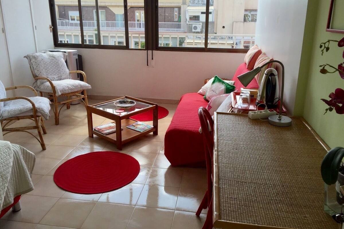 A living room with a red sofa, a wooden coffee table, and a chair. There is a window with a view of buildings outside.