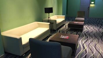 A hotel room with a sofa, armchair, and a small table with a magazine. There is a lamp and a door in the background.