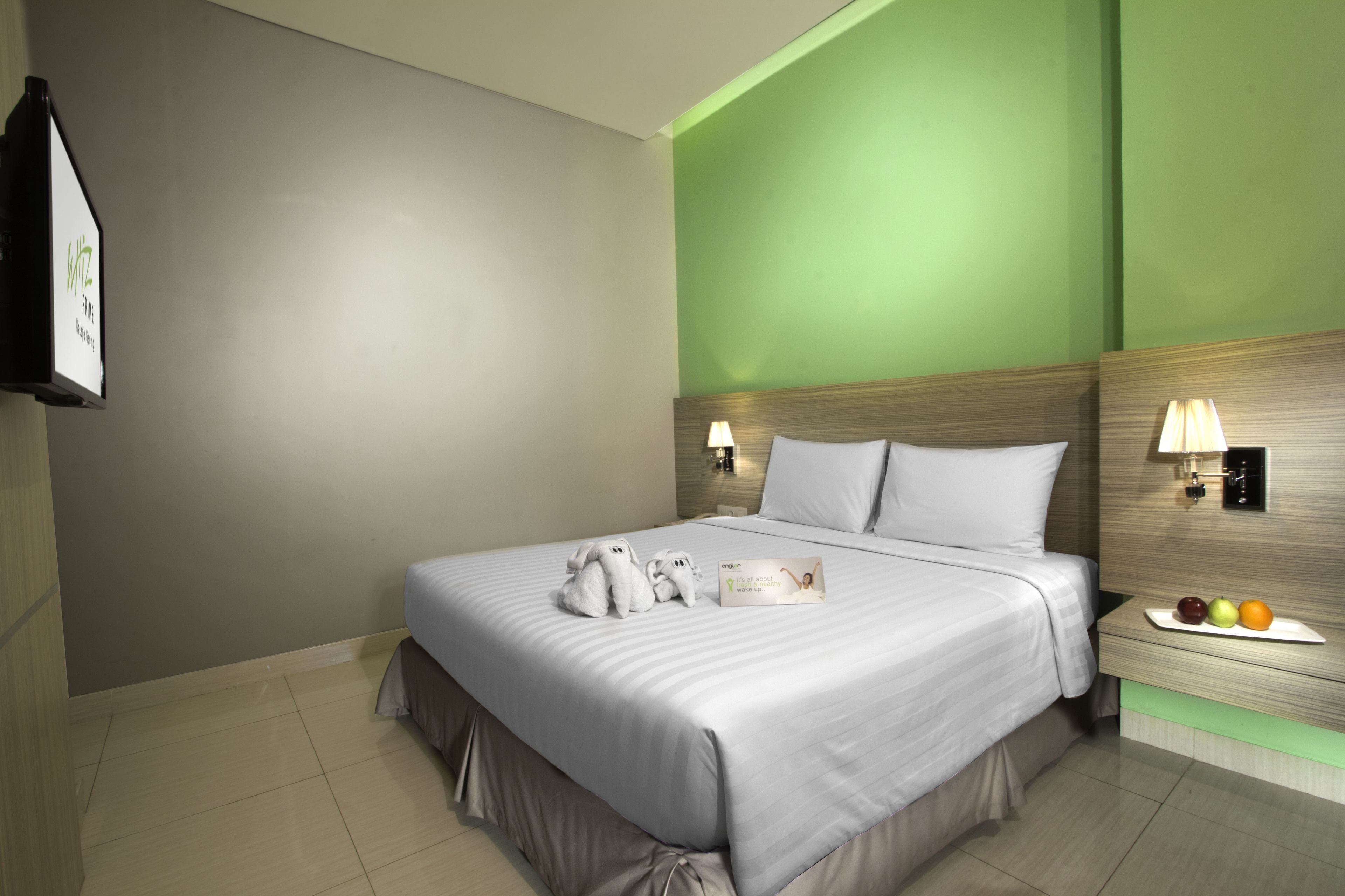 Cama bem arrumada com lençóis brancos, mesa de cabeceira com frutas e luminárias fixadas na parede.