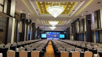 A conference room with rows of tables and chairs, a large chandelier, and a stage with a screen at the front.