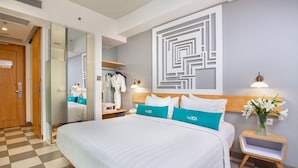 A hotel room with a large bed, a wardrobe, a mirror, a bedside table with a lamp, and a wall with a geometric design. - THE 1O1 Bogor Suryakancana (Bogor)