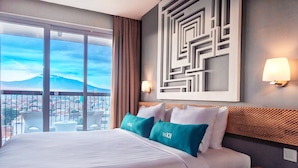 A hotel room with a large bed, two pillows, a nightstand, a wall-mounted lamp, and a view of a cityscape and mountain through large windows. - THE 1O1 Bogor Suryakancana (Bogor)