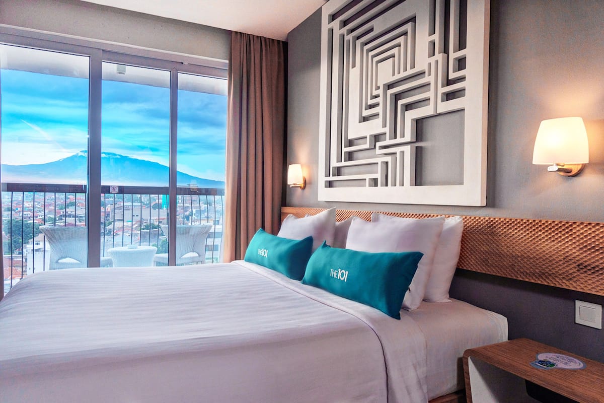 A hotel room with a large bed, two pillows, a nightstand, a wall-mounted lamp, and a view of a cityscape and mountain through large windows.